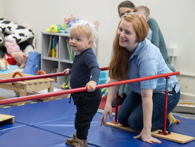 A child practising standing between parallel bars with support from the Little Discoverers team.